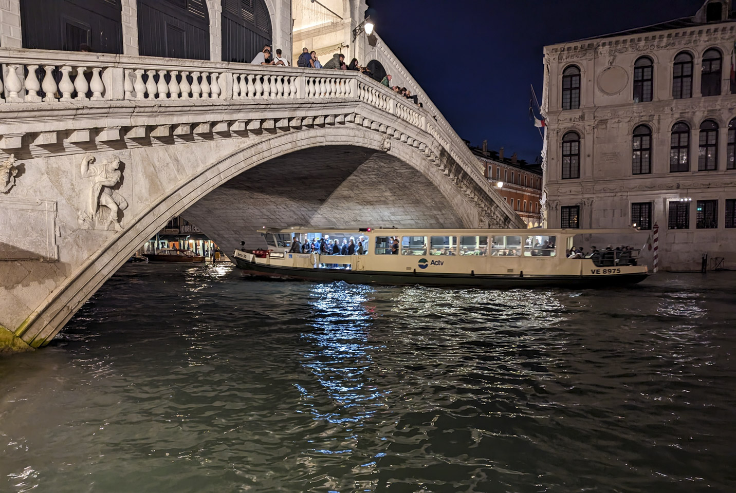 A view of the Rialto bridge at night, with a long motor boat going underneath it. There are people standing and sitting on the waterbus.
