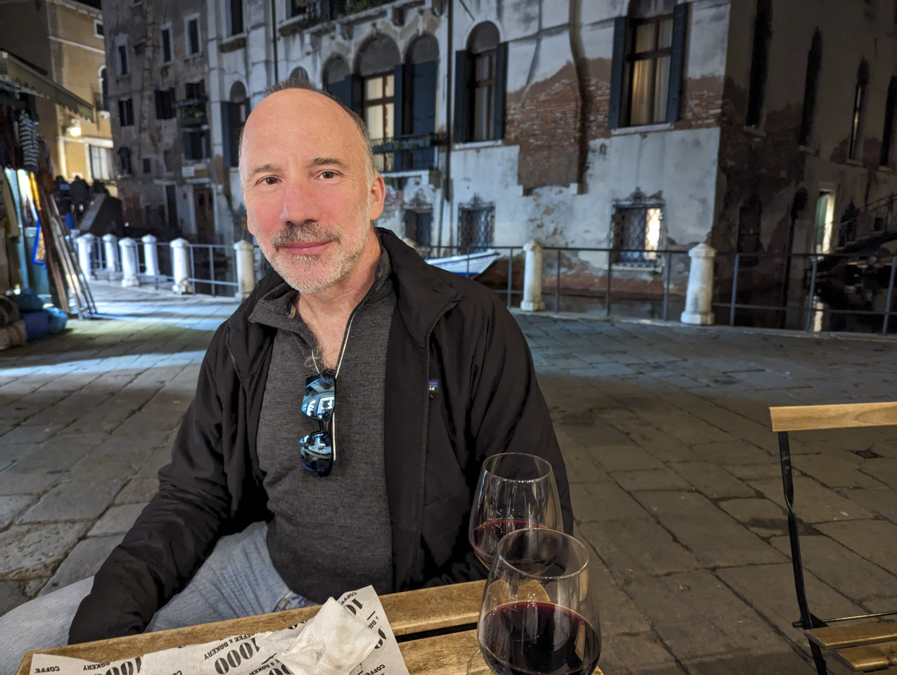 Paul is sitting at an outside table, with a glass of wine in front of him, and some food wrappers.