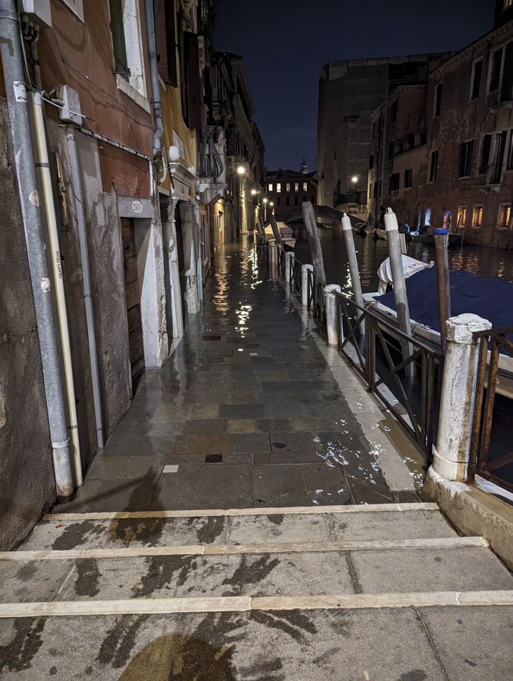 In the early night, looking down a walkway on the side of a Venetian canal, the walkway is covered with a layer of water. There are wet marks on the steps near us, where people have stepped out of the water and on to the dry step.