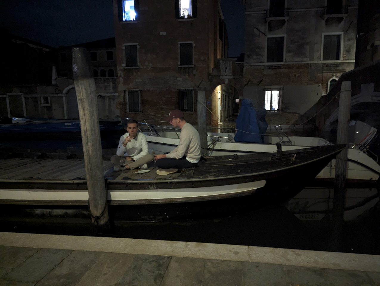 A boat with a wooden top floats in a canal, while two young men sit cross-legged on it while eating some snacks. A glass goblet with wine is in front of one of the men.