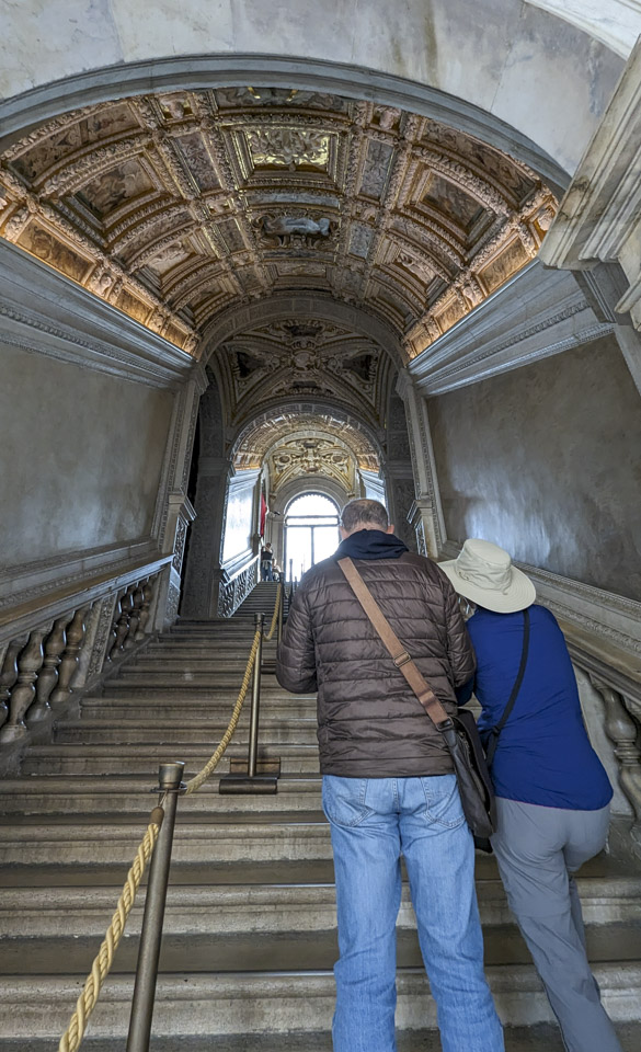 A large staircase with two people looking away from us. The ceiling of the stairwell is extremely ornate with recessed sculpted panels.