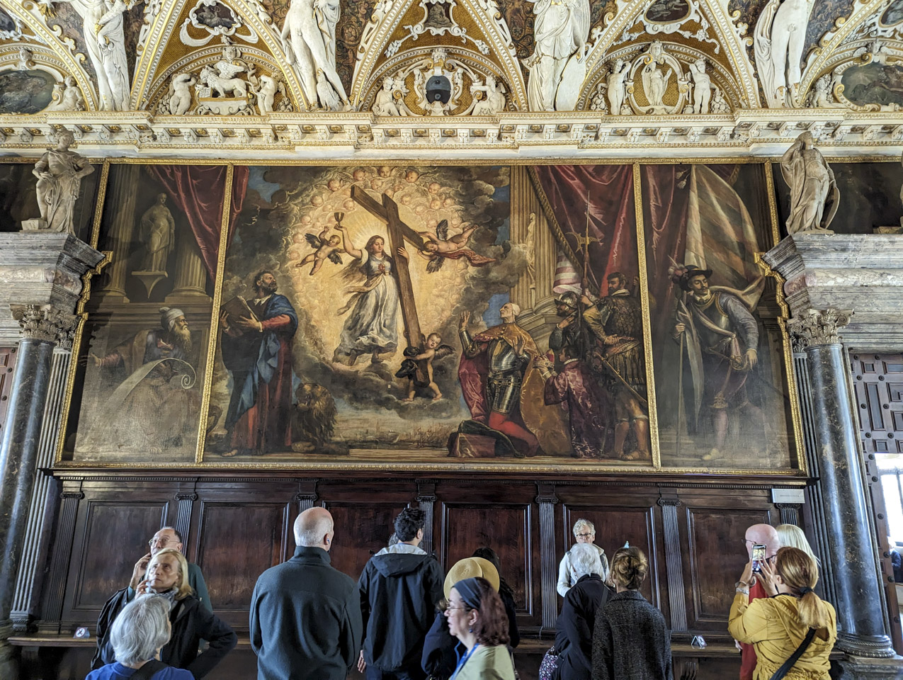 People standing and looking at a large painting on the wall; the painting has a man in armor kneeling, and a woman with light radiating from her while she is holding a cross and a chalice.