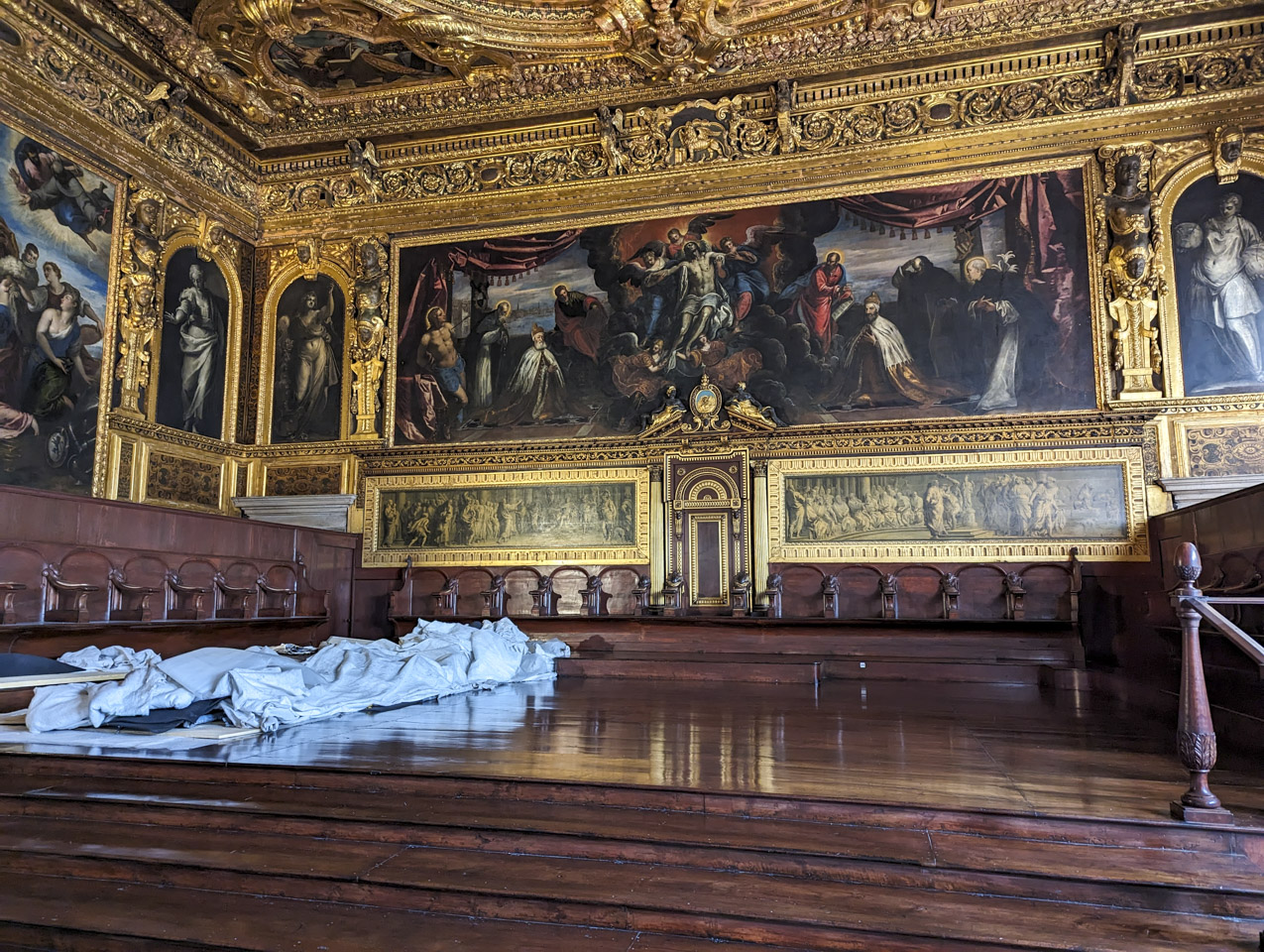 The front of the room, with rich wooden flooring and the same ornate gold carved wall and ceiling. The large painting on the wall shows Jesus being lifted by angels.