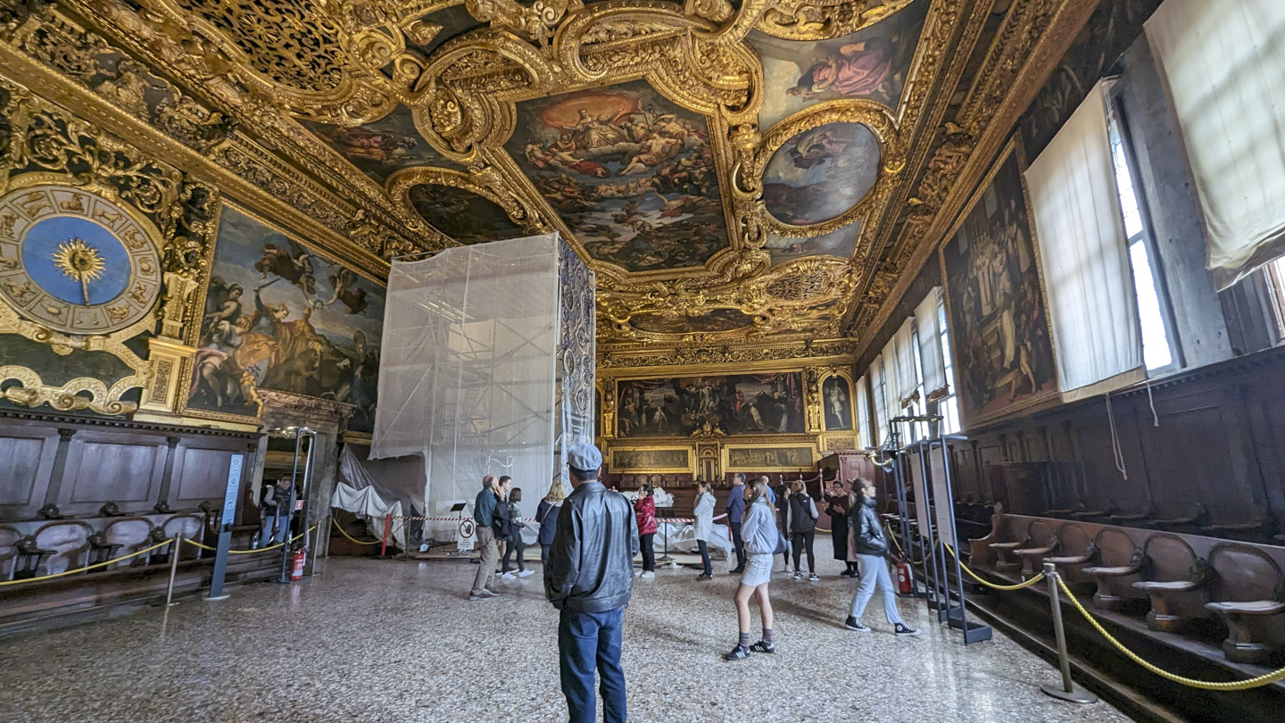 Photo of a large room with built-in seating around the edges and tourists standing in the center. The walls and ceiling are ornately sculpted and gold colored, and there are painted panels recessed in them. A large scaffold covered with mesh walls is in the far section of the room.