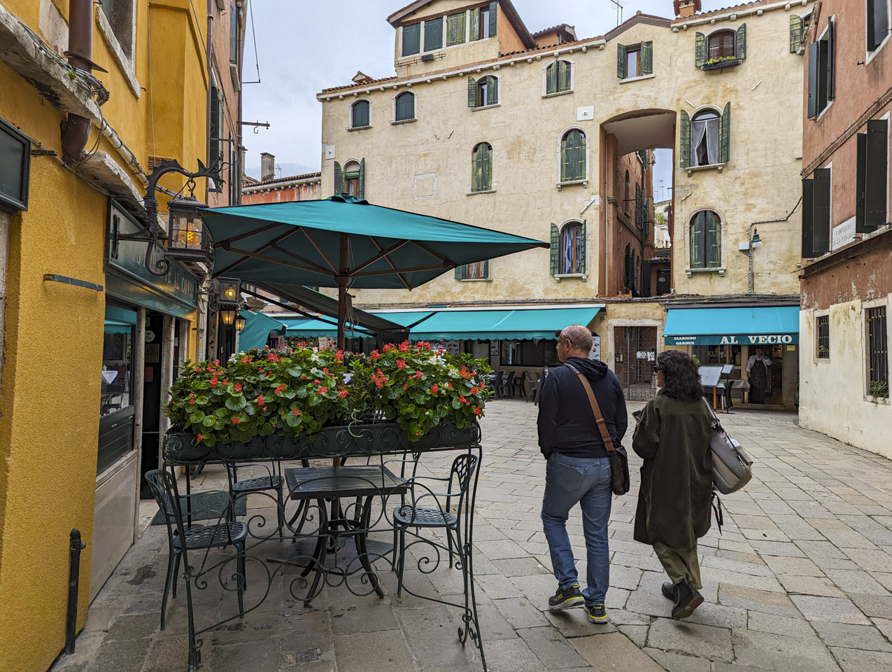 Francesco and Doni walking into a small plaza, looking for the restaurant. There are many green awnings over tables for diners.