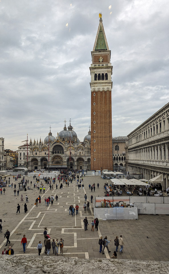 View from the east end of St. Mark's Square. The bell tower and the basilica are visible at the far end. The left side building cannot be seen, but the right side building is visible and the canopies of restaurants are up in the plaza itself.
