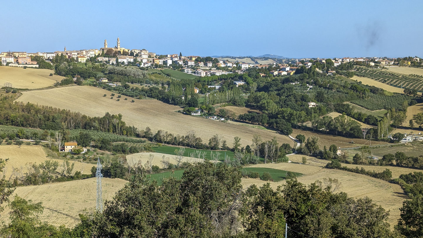 In the foreground are fields and trees, including olive trees. The buildings of the village run in a line on the crest of a low ridge.