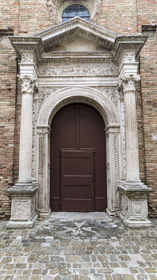 A white stone portal around a large reddish-brown door. The stone has intricate carving on it.