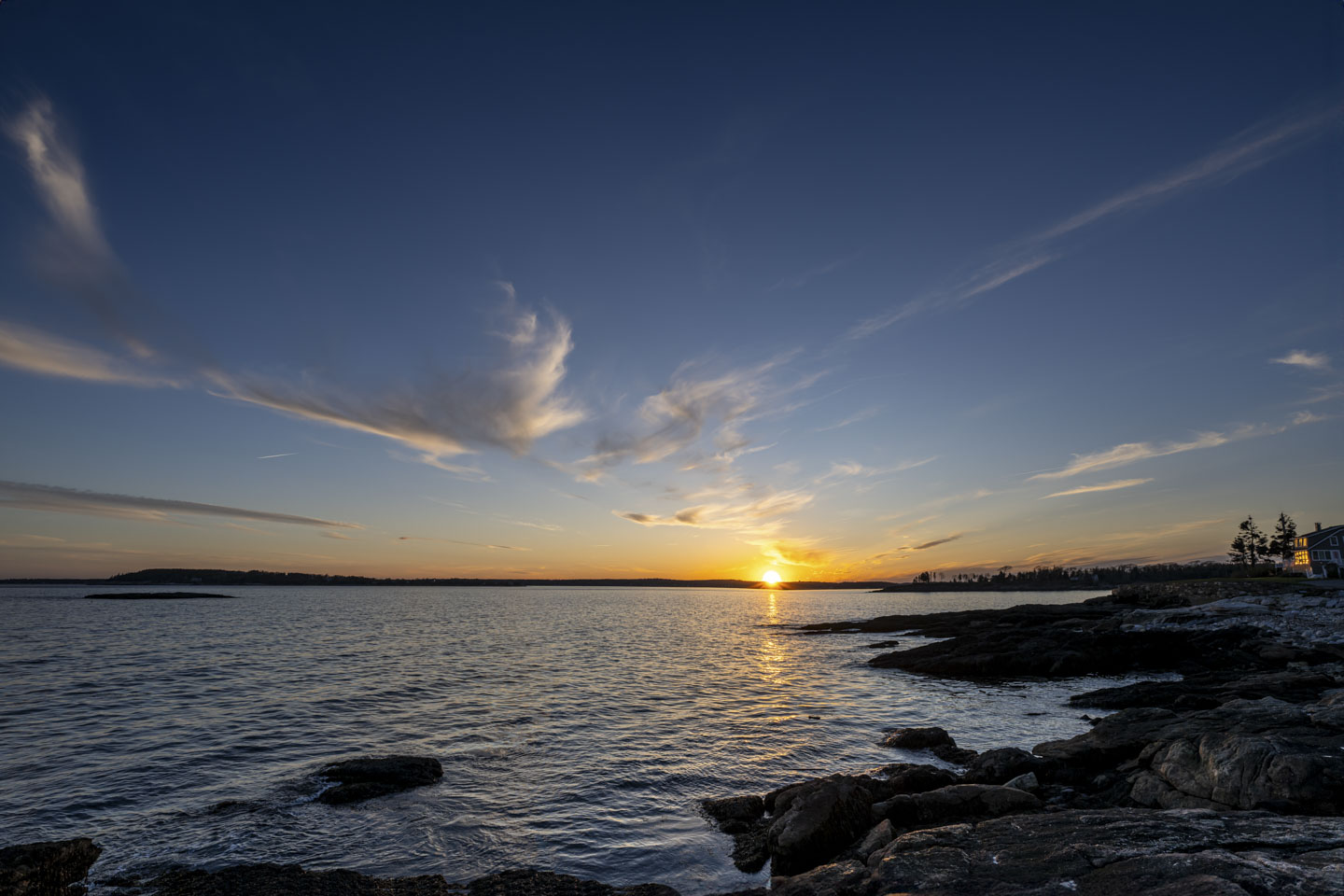 An expansive sky over water and a rocky shore, with the sun setting over land on the opposite side.
