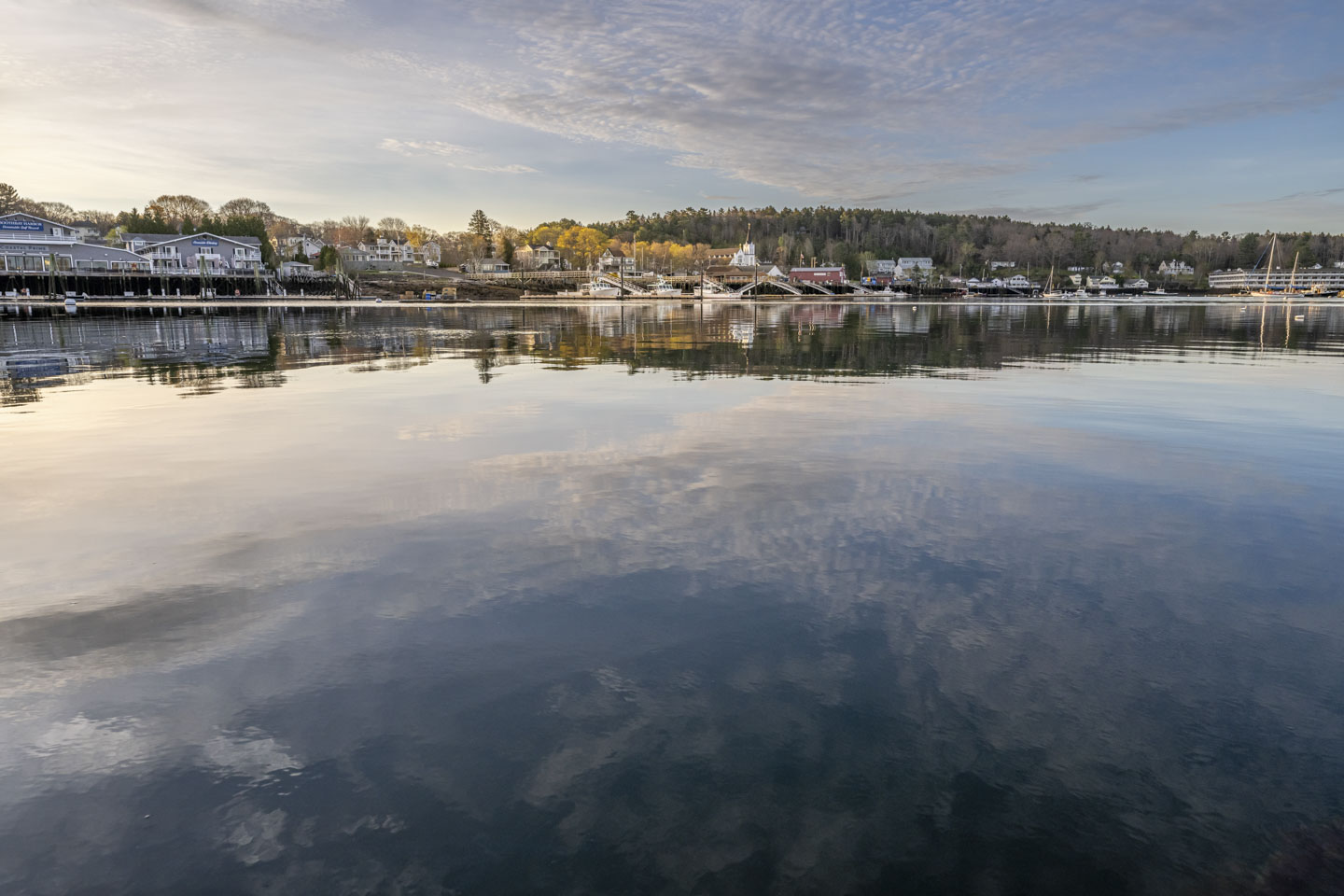 Calm water reflecting clouds. On the opposite shore are buildings, piers, and some trees with colorful spring foliage.