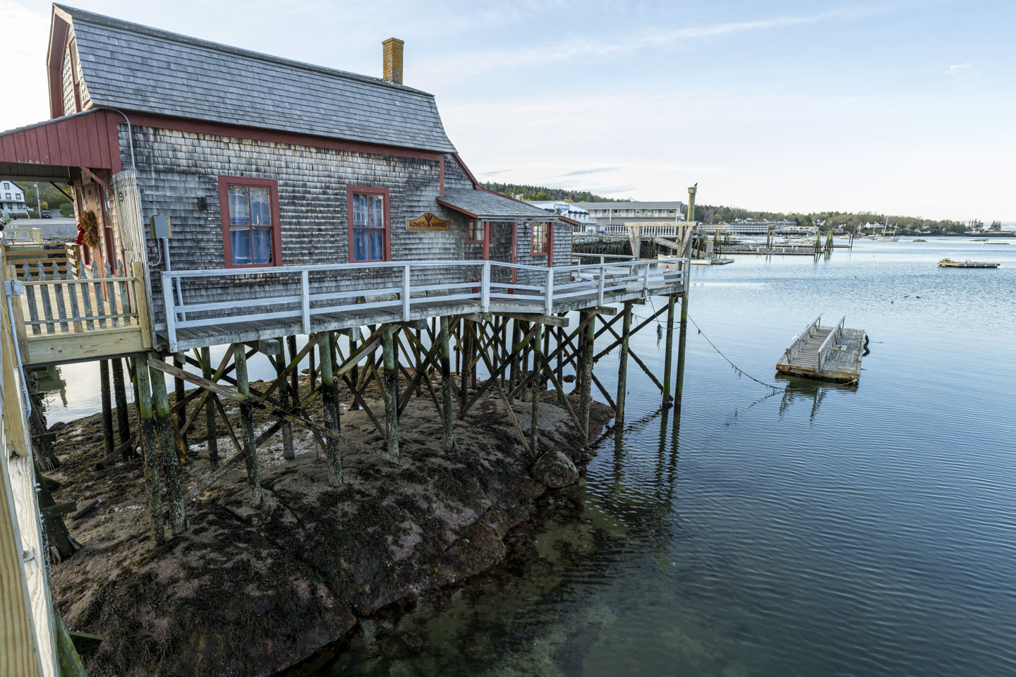 The Bridge House in Boothbay Harbor is seen at low tide. Many wood pilings go to some rock directly below the building, which is visible since the harbor water is low.