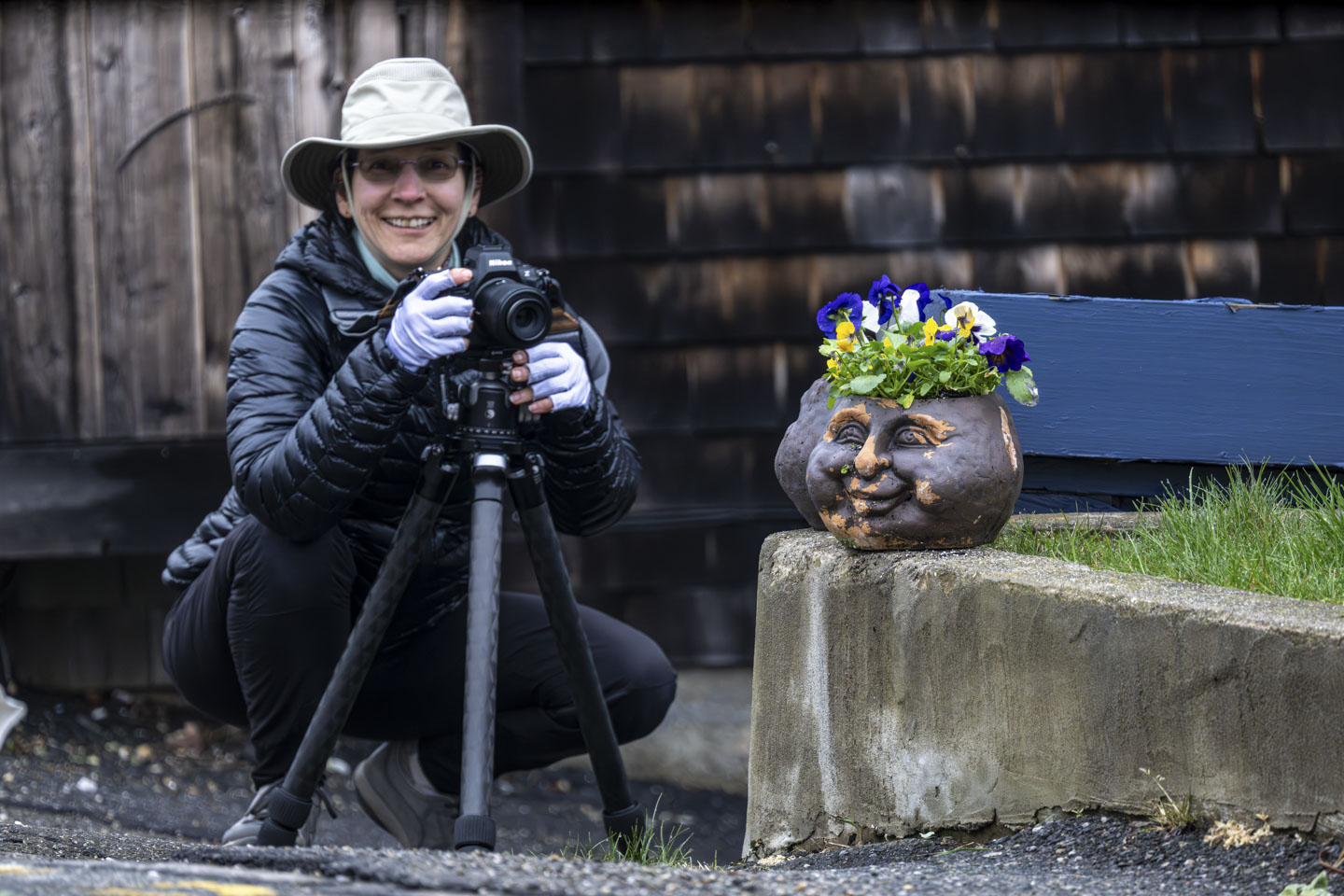 Anne is crouching on the ground holding her camera on a tripod. In front of her are a couple of planters on a low cement wall.