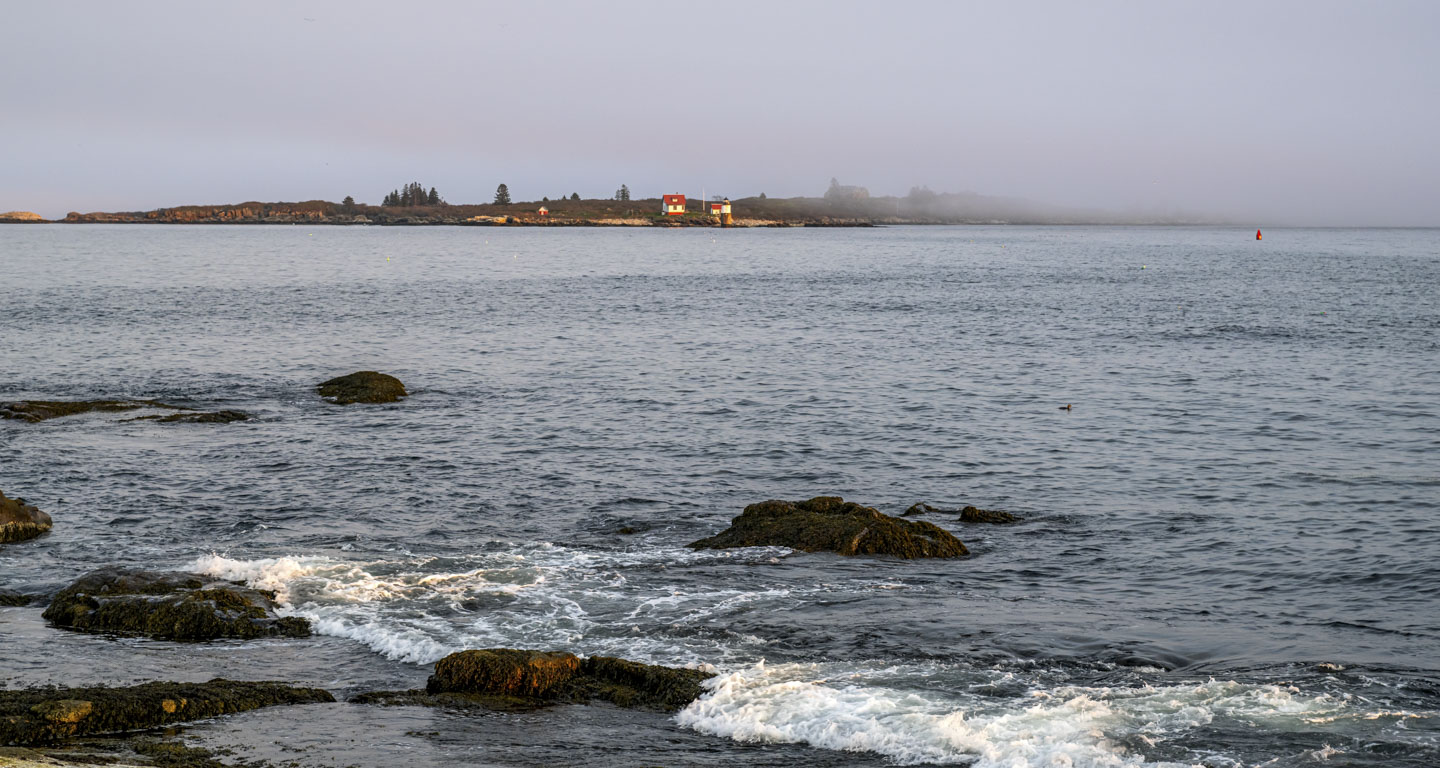 Past some water breaking lightly on small rocks, and then more open water, a lighthouse and a small house can be seen.  Fog obscures another building on the island.