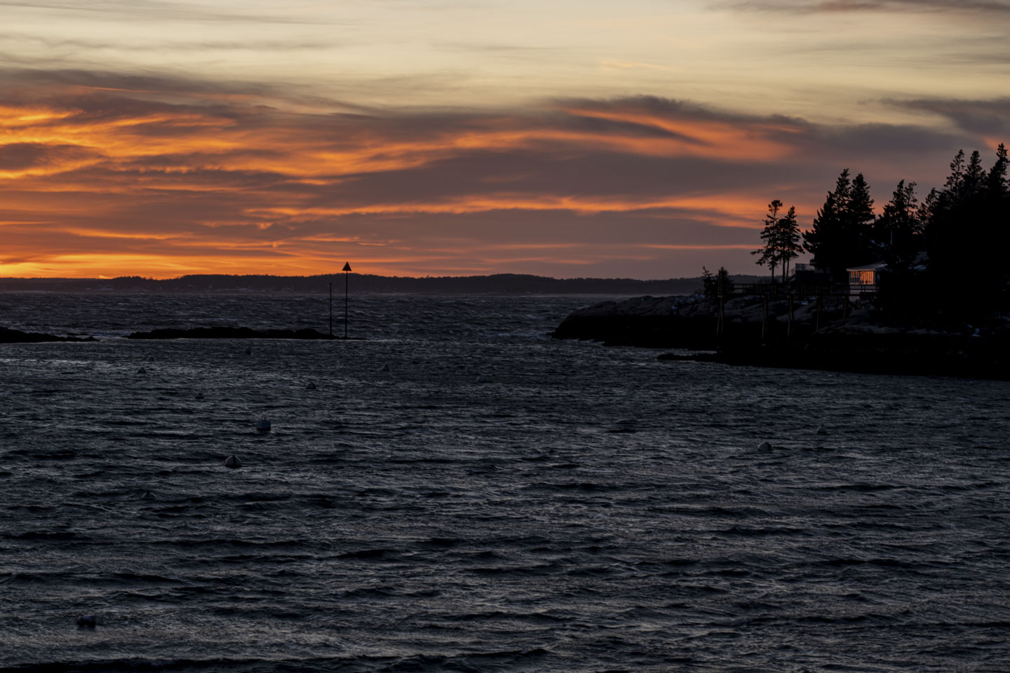 The setting sun, on the left, reflects on the glass of a house on the right. The sky is orange and the water is dark.