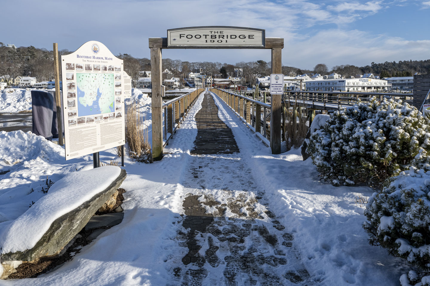 The entrance to the Footbridge in Boothbay Harbor has snow on evergreen bushes and a large rock.