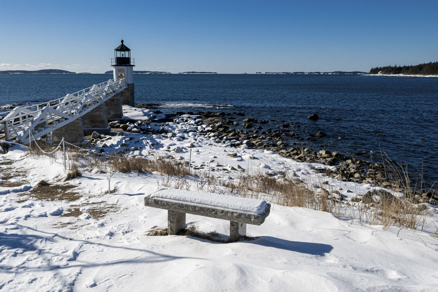 Marshall Point Lighthouse is seen with snow in the rocks, and snow on a bench.