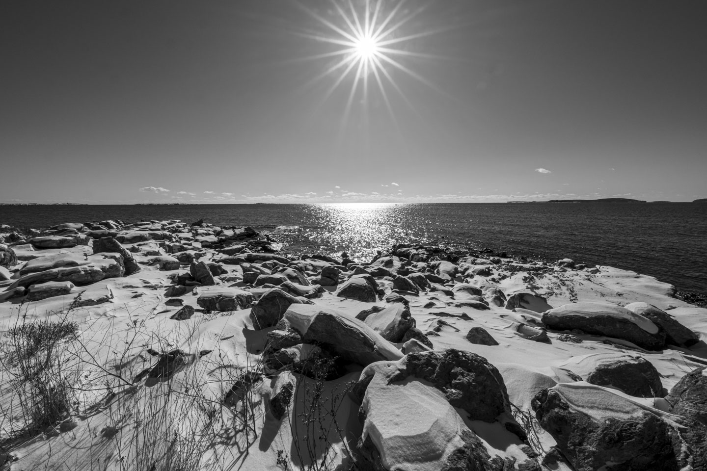 This is a black and white photograph of the sun, with starbursts, above the sparkling ocean and snow-covered rocks.