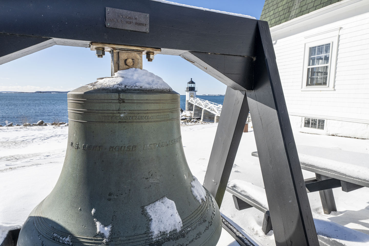 An old very large bell is held by a wooden frame, and the lighthouse can be seen in the distance behind it.