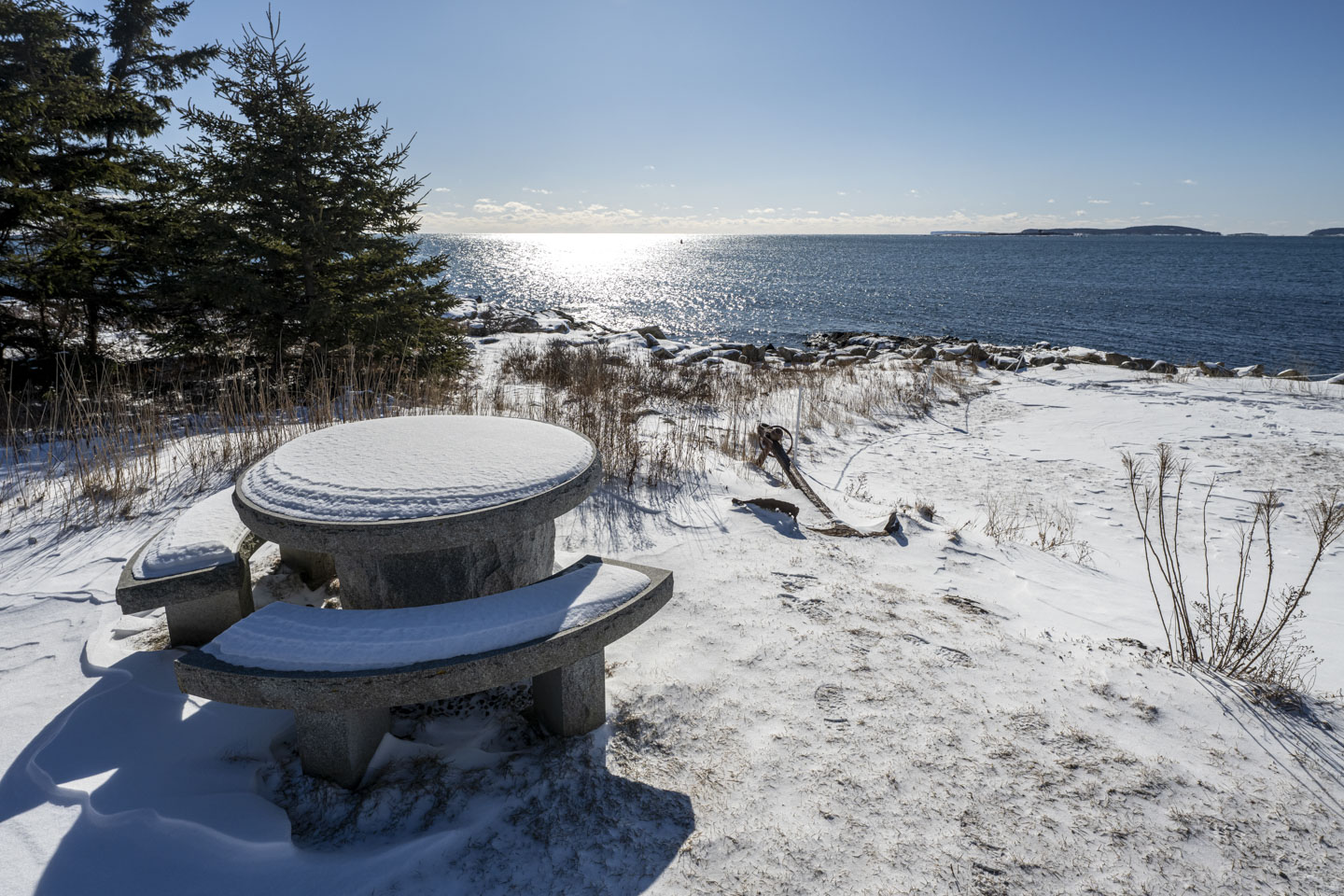 A round cement table with benches has snow on it, and an old anchor is on the ground and partially covered with snow.  The sky is clear and the sun is shining on the water.