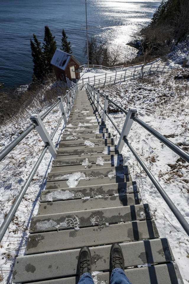A pair of boots are visible at the top of a steep set of stairs that have some snow on them. The sun is reflecting off water in the distance.