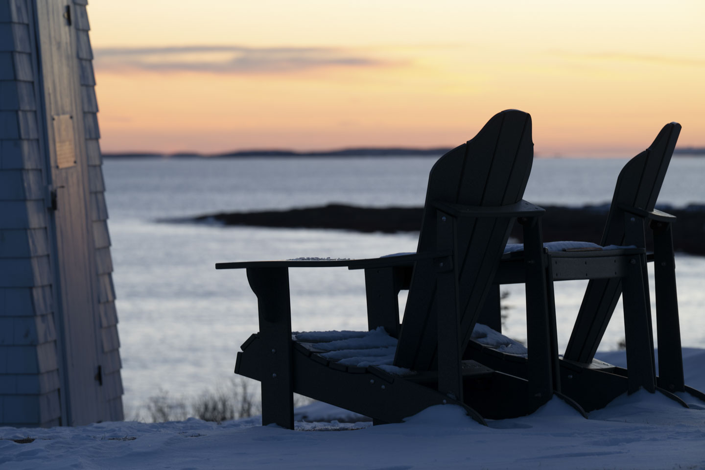 The colors of sunset can be seen in the distance, past two Adirondak chairs that have snow on them.