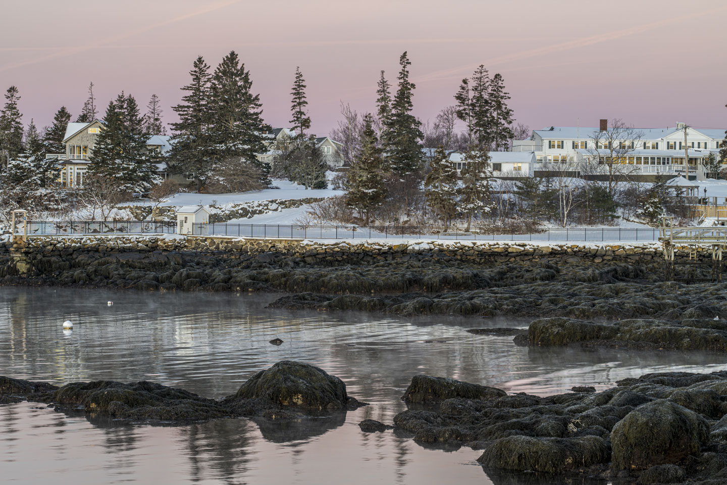A large inn building is on the right, and smaller buildings can be seen among trees on the left. Snow is covering the ground. There is water in the foreground.