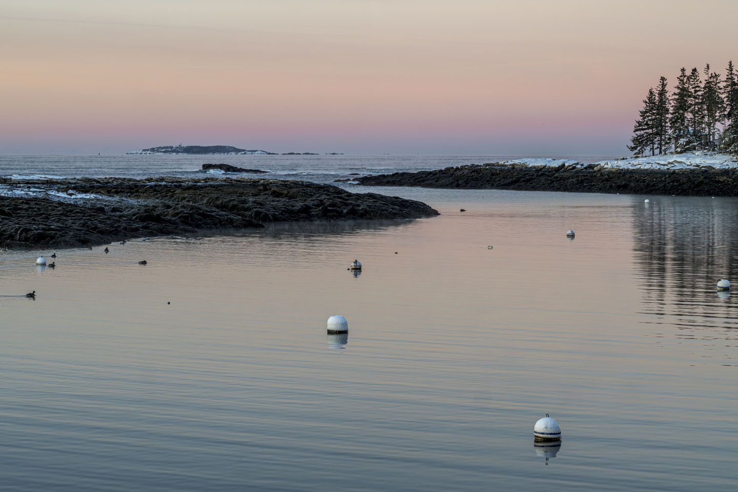 A soft pink sky is reflected in the water, and in the distance a lighthouse on a tall island can be seen.