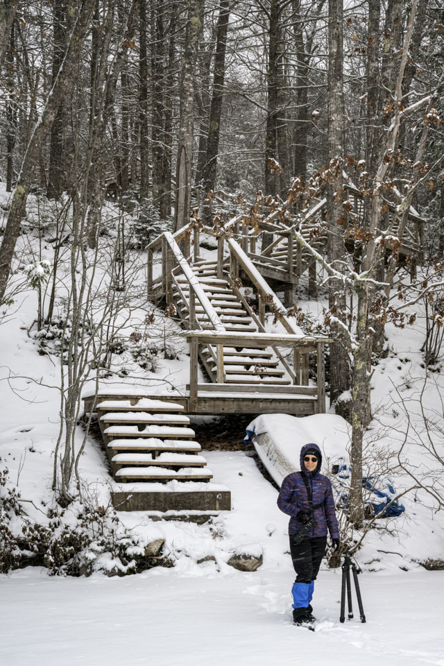 Anne is standing in front of the stairs that go up the hill. She is wearing winter clothing, and snow covers the ground and the frozen lake.