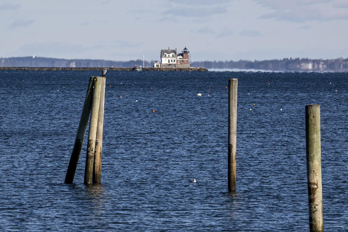 The lighthouse, which is red brick and has a white house attached to is, is seen past 3 mooring posts.