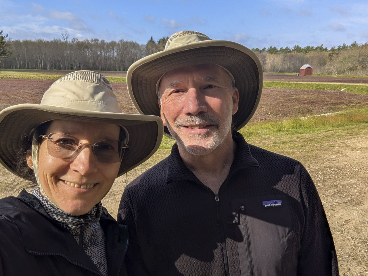Selfie of two people in wide-brimmed hats with the cranberry bog and a small red shed behind them