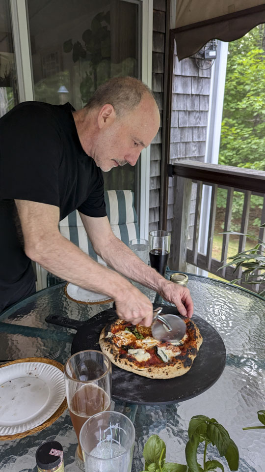 Paul is cutting a delicious looking pizza, which is on a table on the deck.
