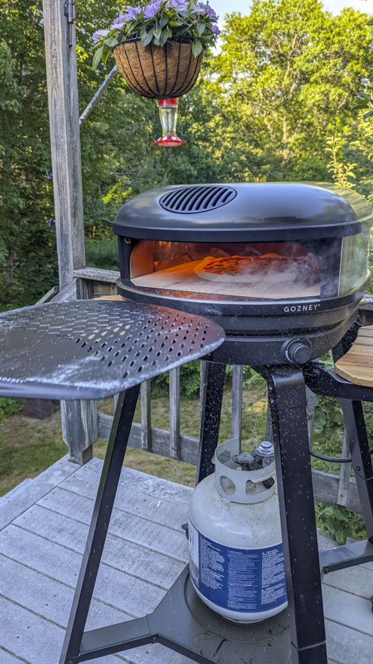 A large black peel is being pulled away from the pizza oven, with the flame glowing inside the oven.