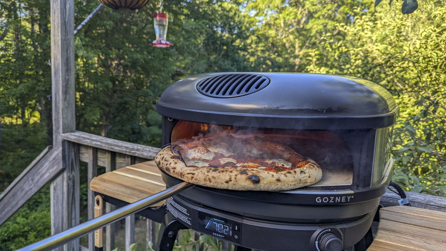 The turning peel is holding a steaming pizza in the mouth of the pizza oven.
