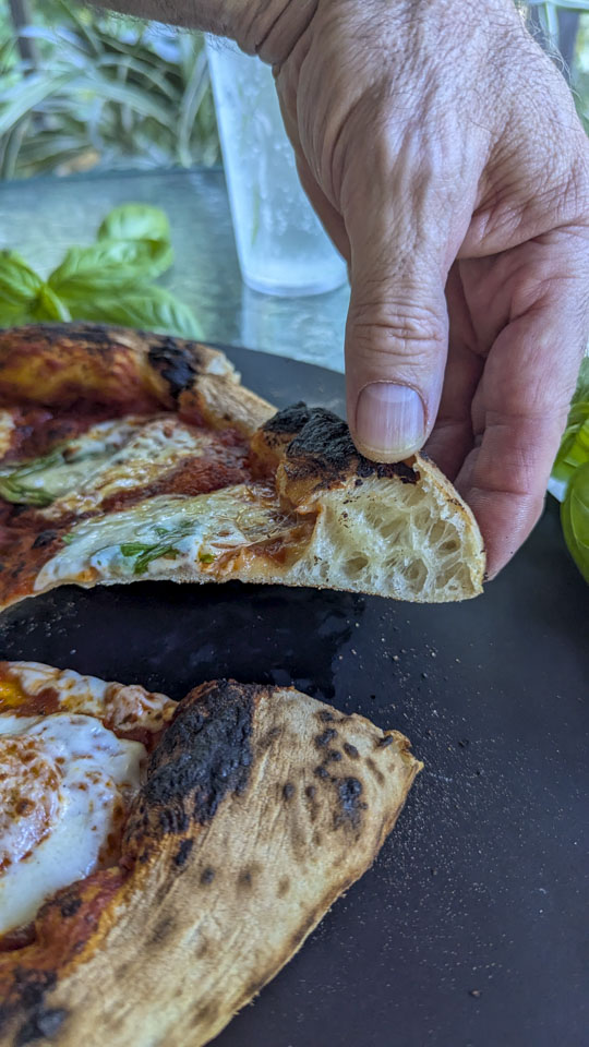 Paul's hand is holding up a cut slice of pizza, showing the crust.