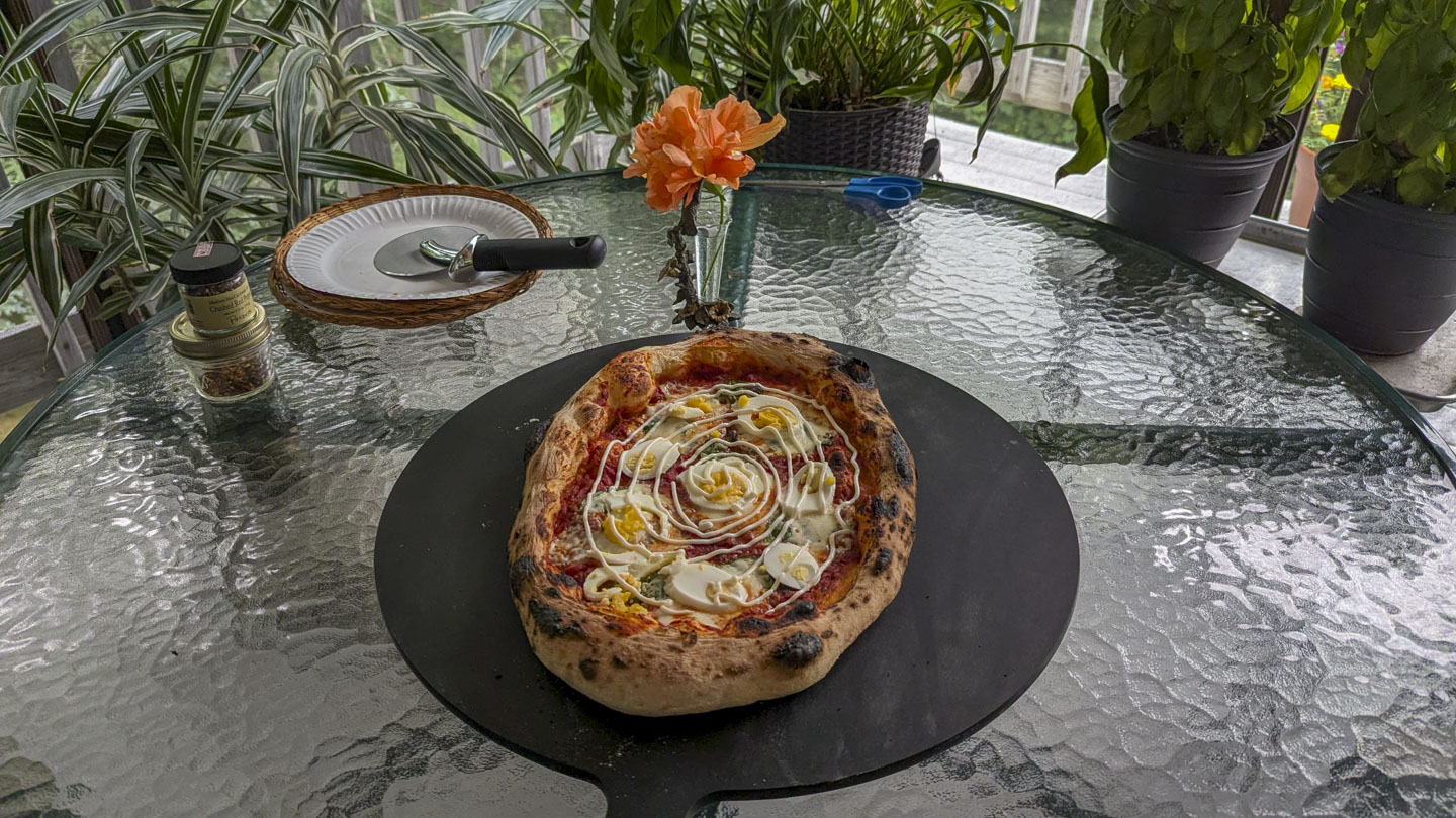 Pizza Rossini on a black cutting board on a glass table, with plants and a flower around it.