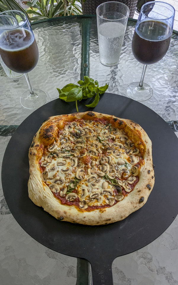 A nicely formed mushroom pizza, with 2 goblets of beer and some basil.