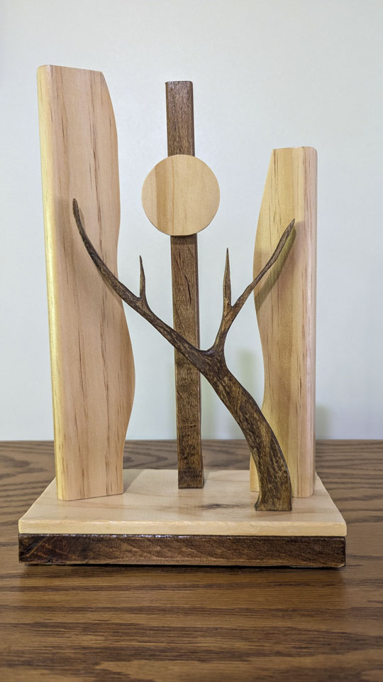 Wooden sculpture of a tree branch in front of 3 upright columns, two being curved and the center on having a round disk representing the moon.